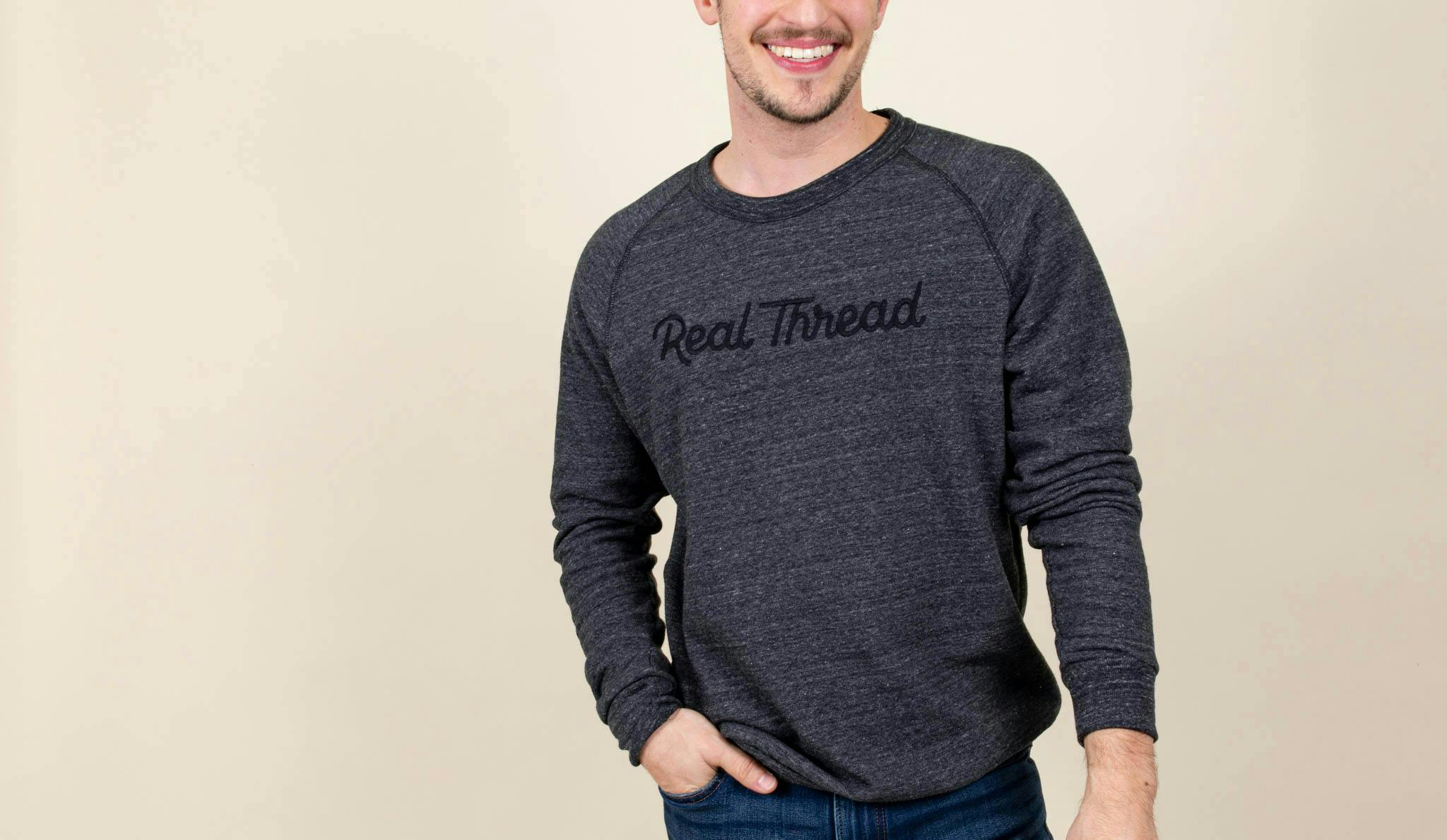 A man wears a heathered black sweatshirt with a black Real Thread embroidered on the front 